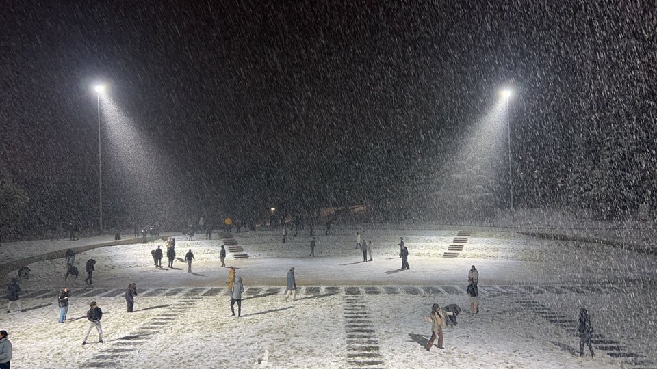 Heavy snow falling across a floodlit sports field at night, students scattered across it.