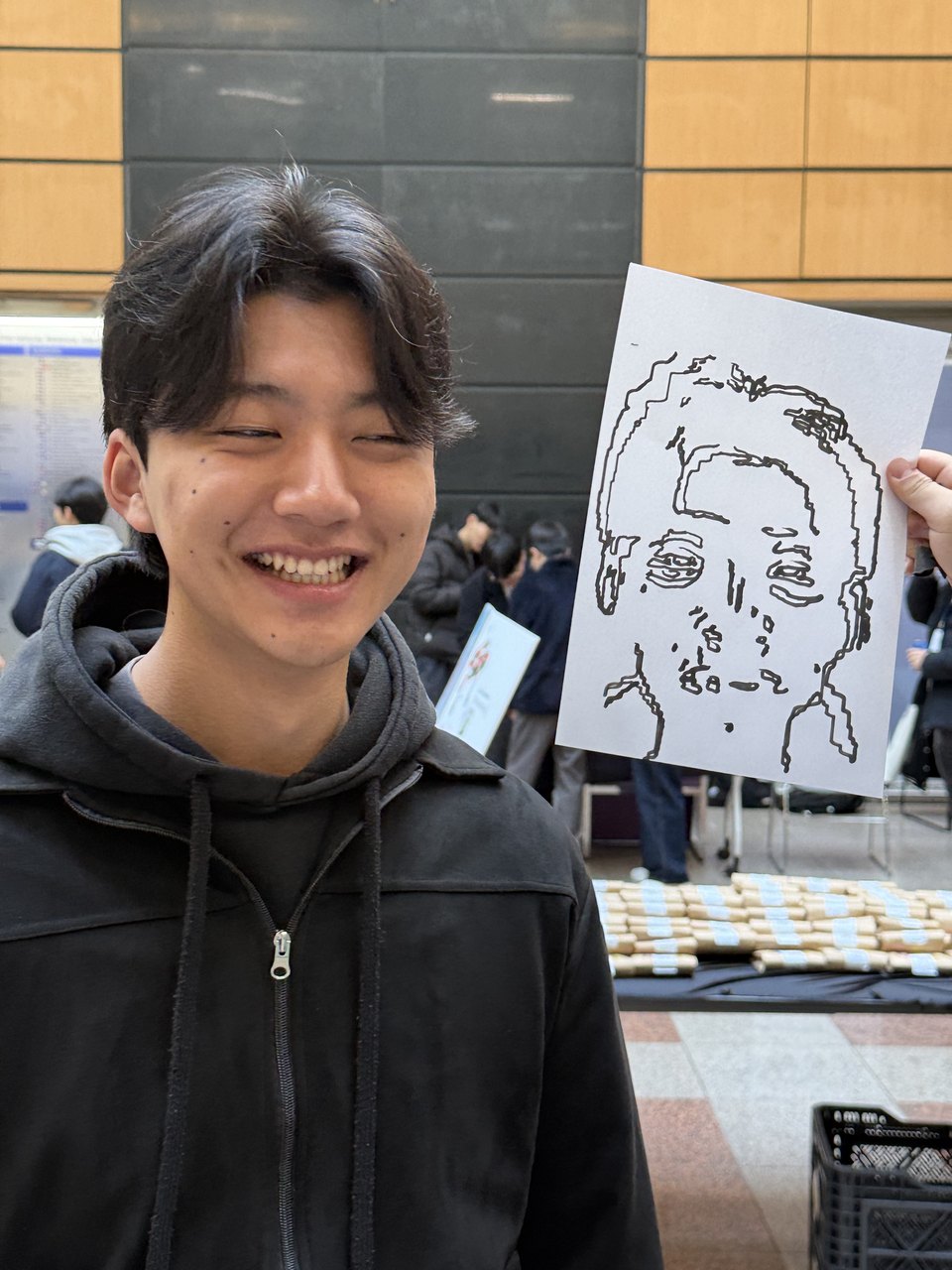 A student smiling and holding up a line-drawn caricature of his own face, drawn by the machine.