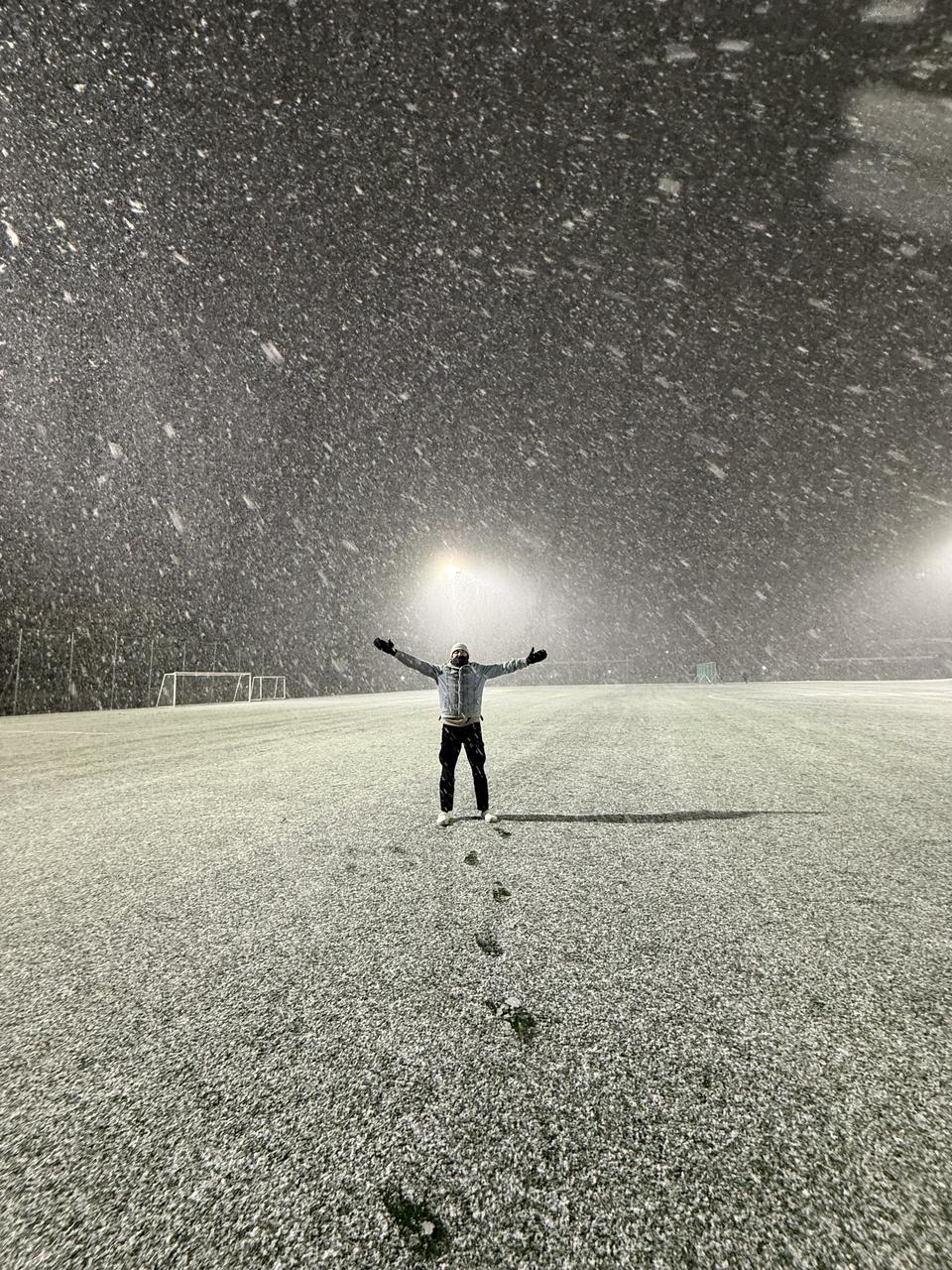 Abdullah alone on a floodlit sports field in heavy snow at night, arms spread wide, his footprints leading back across the pitch.