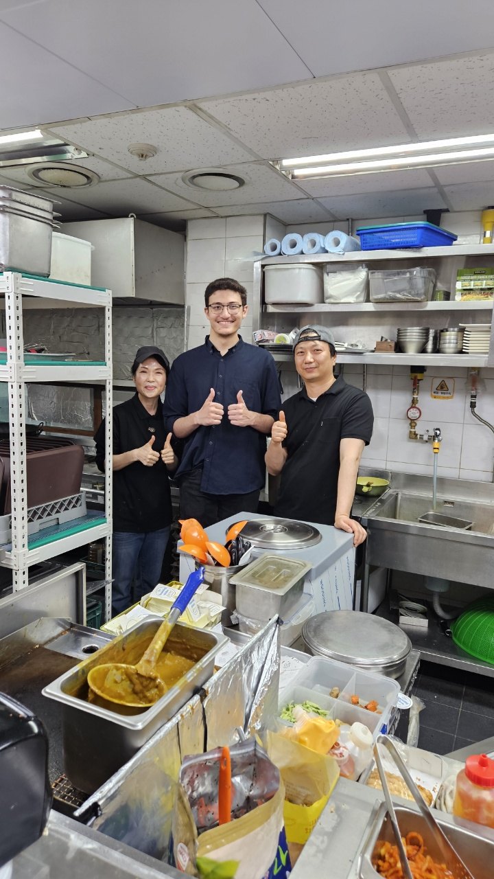 Abdullah in a commercial kitchen with two colleagues, trays of prepped food in front of them.
