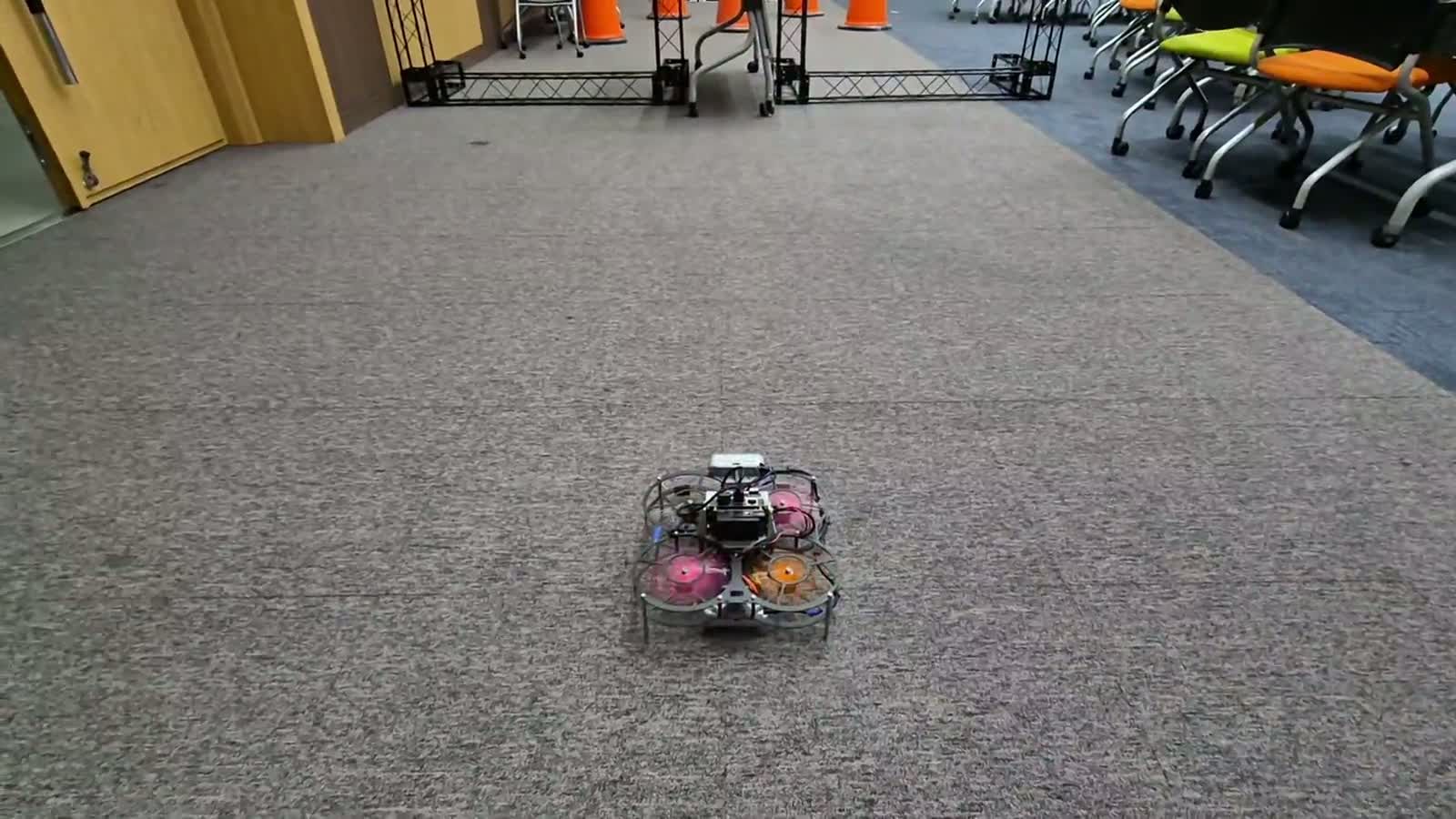 The drone flying the mission course indoors: past traffic cones, through a truss gate marked with AprilTags, towards a row of mock storefronts.